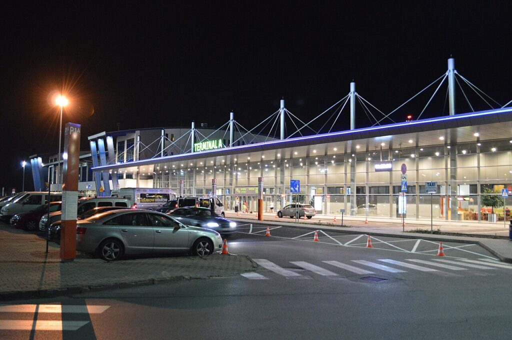 Night exterior of Terminal A airport with a glass facade, neon 'TERMINAL A' sign, and cars parked in front of the terminal.