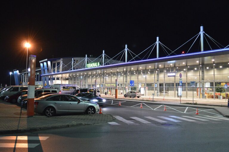 Night exterior of Terminal A airport with a glass facade, neon 'TERMINAL A' sign, and cars parked in front of the terminal.