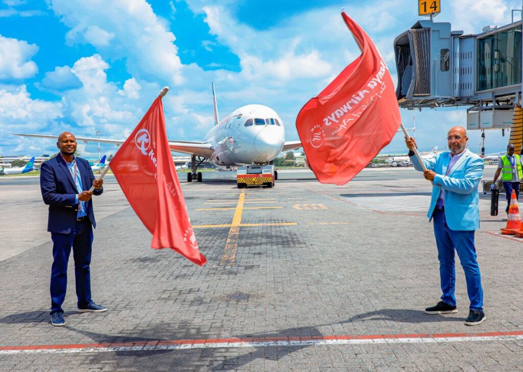 Two men in smart casual outfits wave red flags on an airport tarmac in front of a white passenger jet by the gate.