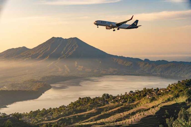 Airplane descending toward a valley with terraced hills, a lake, and a distant volcanic mountain at sunset.