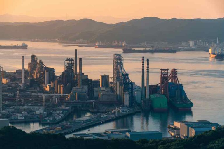 Industrial port at dusk with cranes, smokestacks, and cargo ships on calm water, mountains in the background