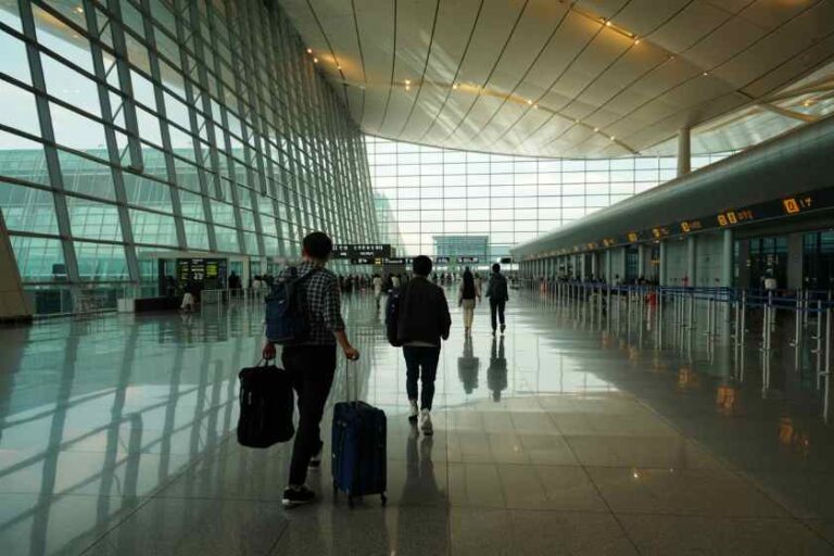 Travelers walking through a modern airport terminal with rolling suitcases under a glass wall ceiling.