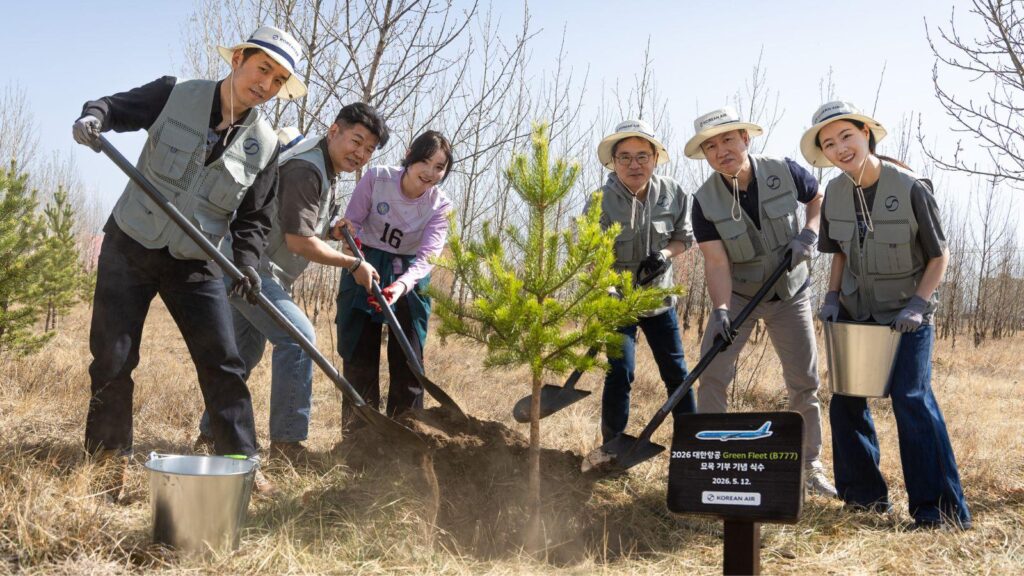Group of volunteers in gray vests plant a young sapling together outdoors, using shovels and metal buckets in a dry field.