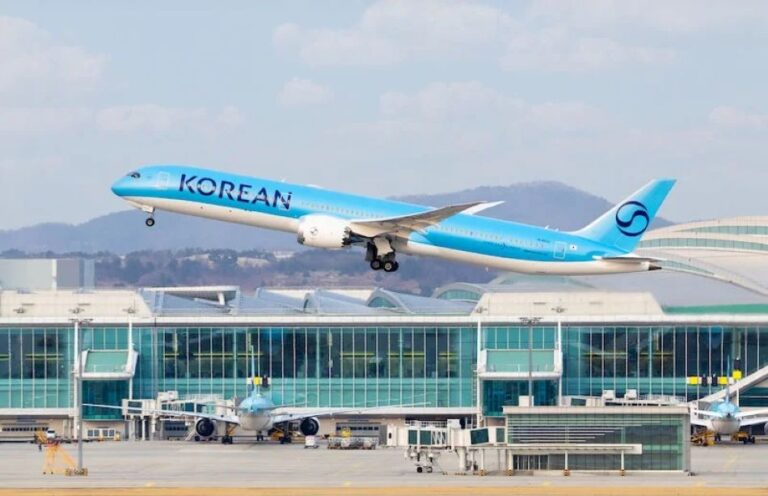 Blue Korean Air jet lifting off from the runway with a glass-terminal airport in the background, hills beyond.
