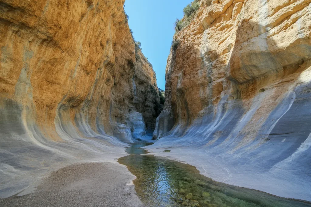 Narrow canyon with orange-brown rock walls and a pale gray lower section; a shallow, clear stream runs between the walls.