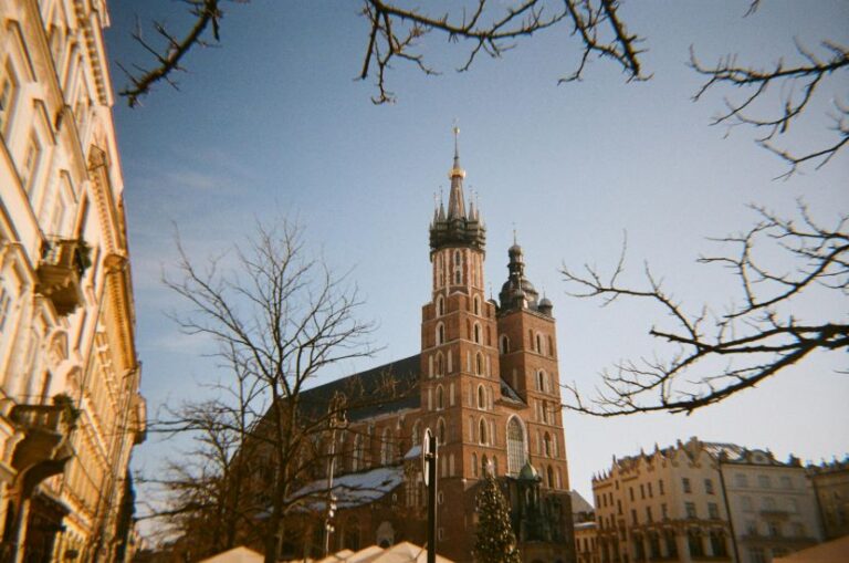 Brick Catholic church with tall spires in a snowy urban plaza, bare trees and surrounding historic buildings against a clear blue sky