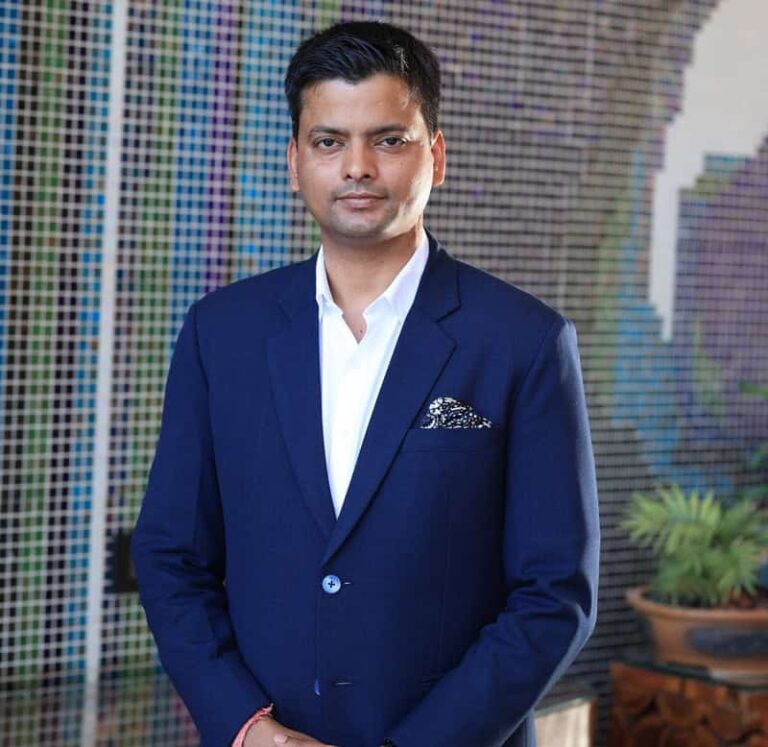 Portrait of a man in a navy suit with a white shirt and pocket square, standing before a colorful mosaic backdrop, posing formally to the camera.