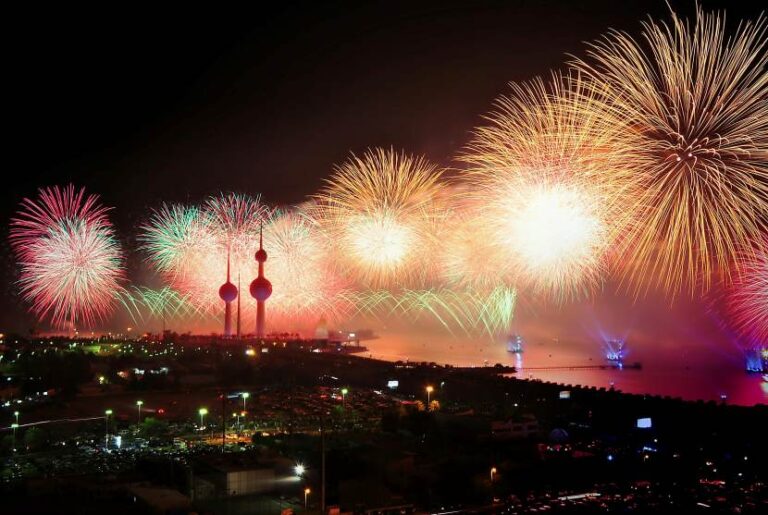 Night view of colorful fireworks over a coastal city harbor, with two tall towers and lit waterfront.