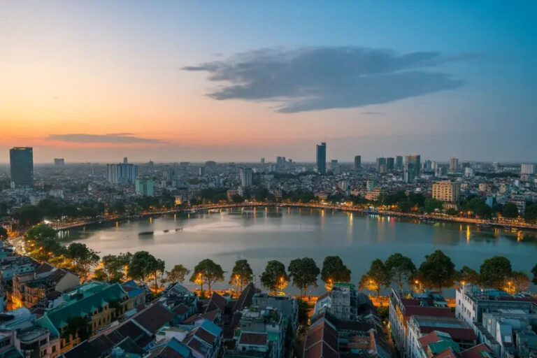 Bird's-eye view of a city at sunset, with a circular lake bordered by trees and a mix of low-rise and high-rise buildings surrounding it.