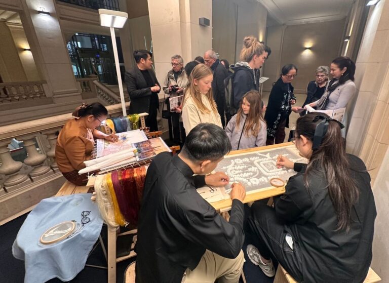 Lacemaking workshop: two artisans weave intricate lace at a table while a group watches in a grand hall.