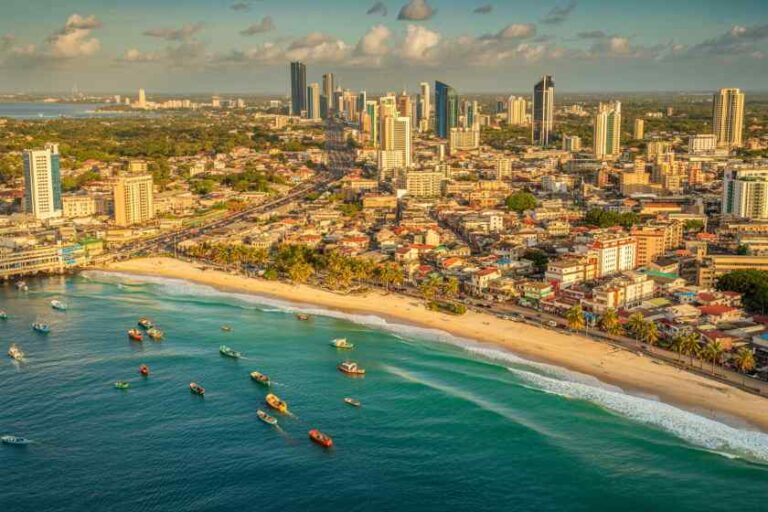 Aerial view of a coastal city with a sandy beach, turquoise water, and a cluster of high-rise buildings along the shore.