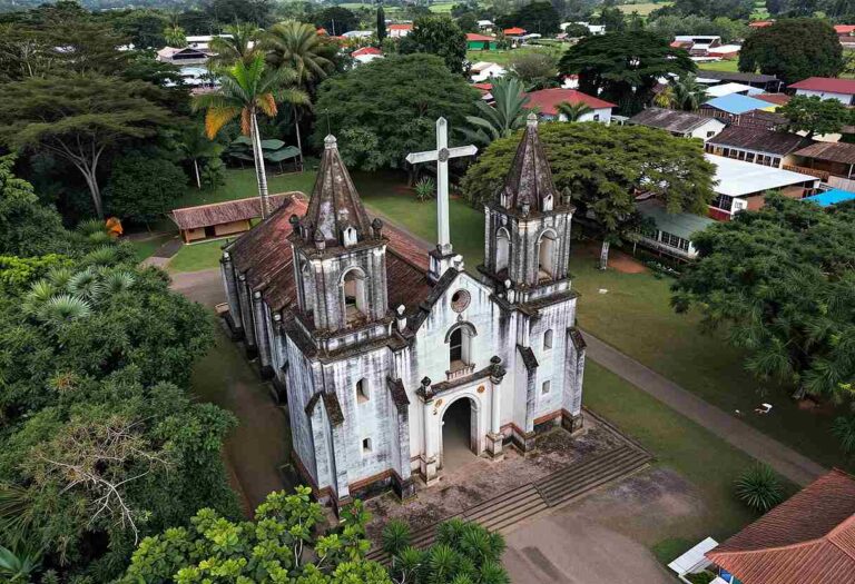 Aerial view of a white church with twin spires and a central cross, surrounded by green trees and a village setting.