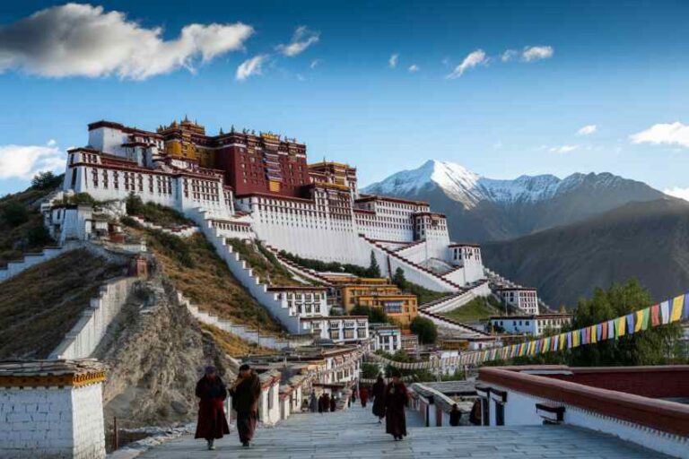 Potala Palace with white walls and red buildings on terraced slopes, prayer flags fluttering, and snow-capped mountains in the distance.