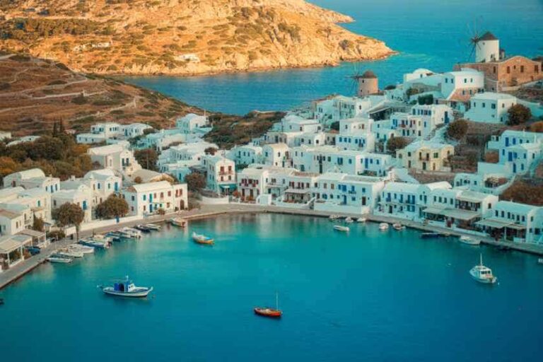 Aerial view of a whitewashed coastal village with blue doors and boats in a calm harbor along turquoise water, hillside townscape in the background.