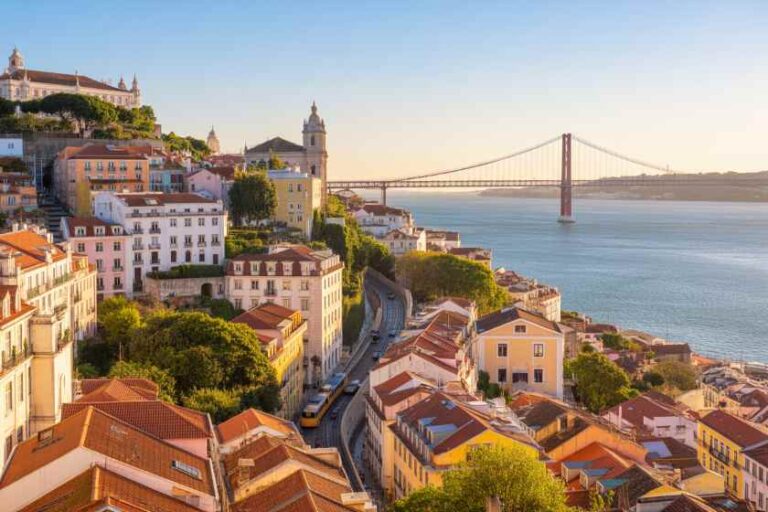 Panoramic view of Lisbon’s Alfama district: colorful hillside buildings, a yellow tram on a winding street, and the Tagus River with the 25 de Abril Bridge in the distance.