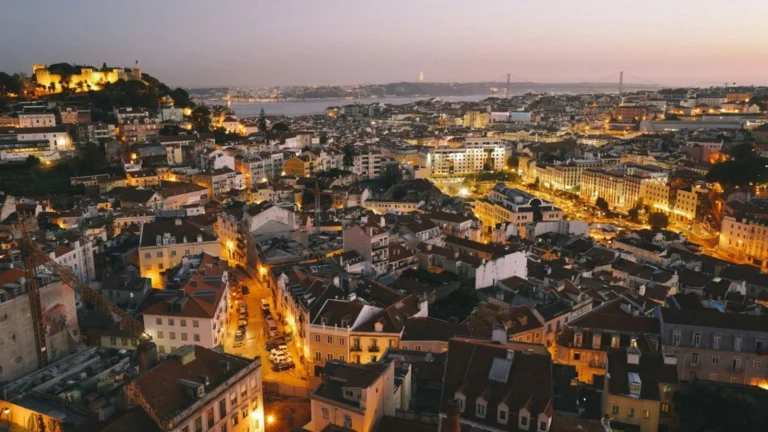 Aerial view of a Mediterranean city at dusk with warm streetlights, a river in the distance, and a bridge across it.
