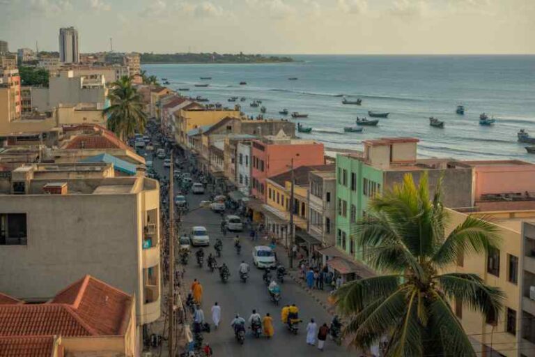 Busy seaside street lined with pastel buildings and palm trees, boats anchored in the calm ocean to the right.