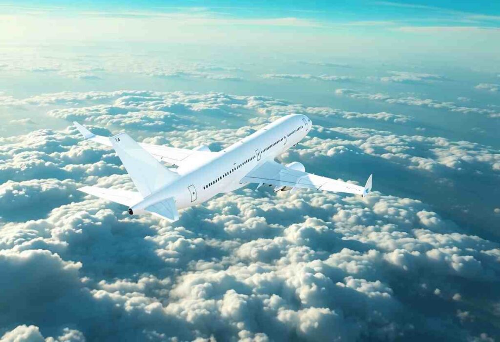 White commercial airliner flying above fluffy clouds in a clear blue sky, viewed from a rear-side angle.