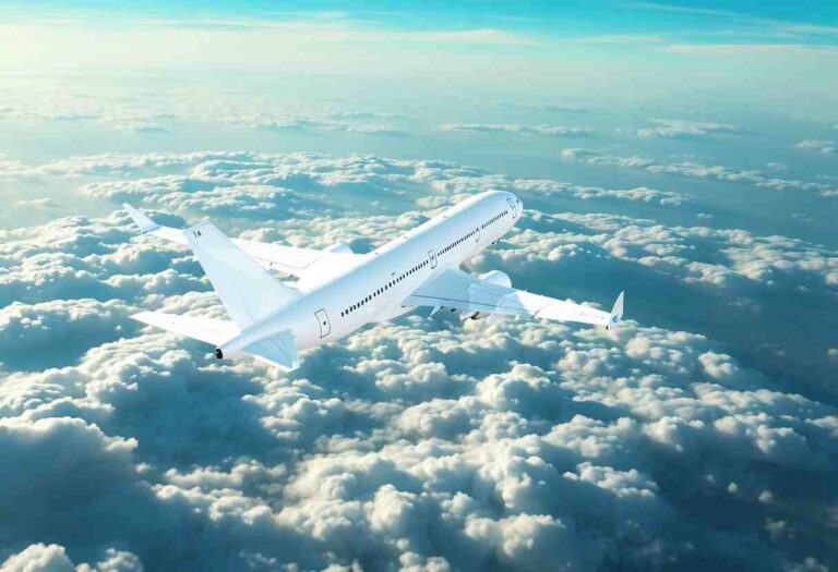 White commercial airliner flying above fluffy clouds in a clear blue sky, viewed from a rear-side angle.