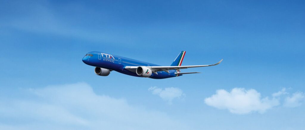 Blue ITA Airways passenger jet in mid-flight against a clear blue sky with a white‑cloud horizon behind it.