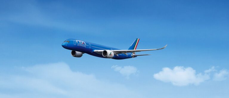 Blue ITA Airways passenger jet in mid-flight against a clear blue sky with a white‑cloud horizon behind it.