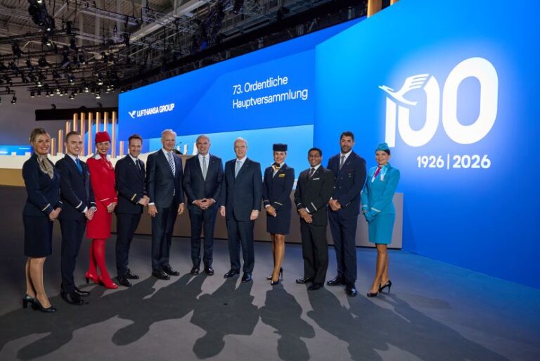 Group of Lufthansa staff and executives in formal uniforms posing for a photo at a 100th‑anniversary event on a blue stage backdrop.