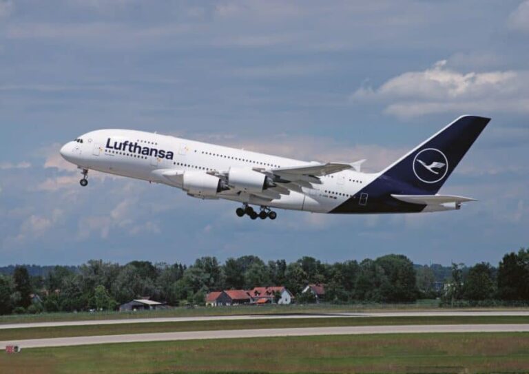 Lufthansa airliner lifting off from a runway with its landing gear down, under a partly cloudy sky and distant trees.