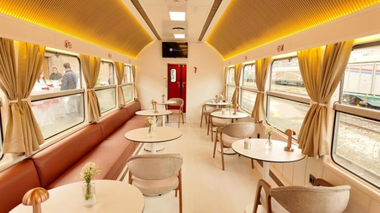 Interior of a train dining car with round tables, beige chairs, and warm yellow lighting along the ceiling.