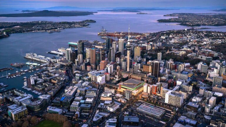 Aerial view of a dense harbor city with tall skyscrapers, including a prominent tower, surrounded by water, ships, and distant green hills.