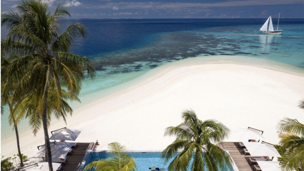 Tropical beach scene with white sand, palm trees, a pool in the foreground, and turquoise sea with a sailboat on the horizon.