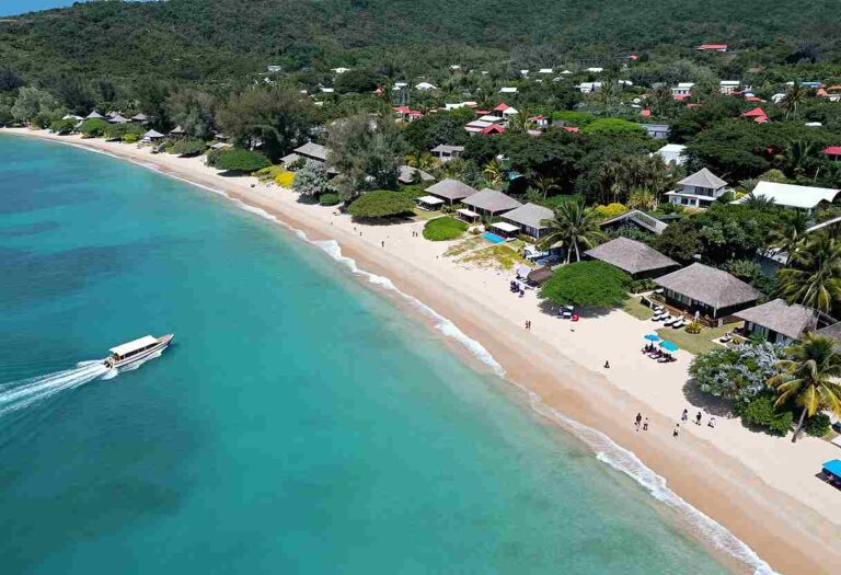 Aerial view of a tropical beach resort with turquoise water, white sand, palm trees, and thatched huts along the shore.