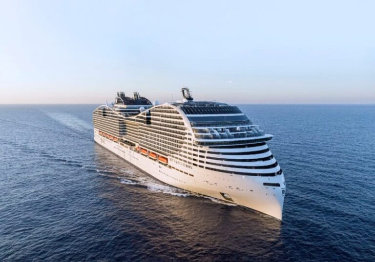 A large white cruise ship sailing on calm blue ocean under a clear sky, viewed from the bow side.