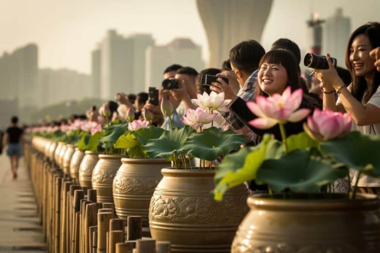 Group of people with cameras along a railing, photographing pink lotus flowers in large ceramic pots with a city skyline in the background.