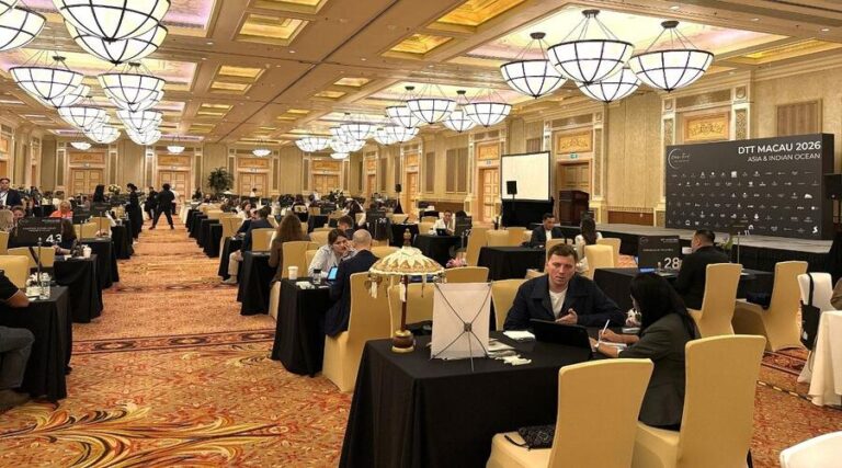 Large banquet-style conference hall with attendees seated at multiple black-table setups, presenting stage in the background, and decorative chandeliers overhead.