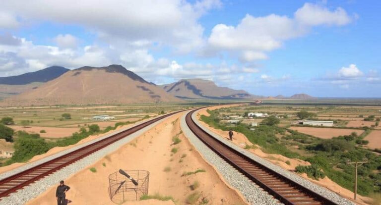 Two railroad tracks diverge across a sandy desert toward distant mountains under a blue sky with clouds.