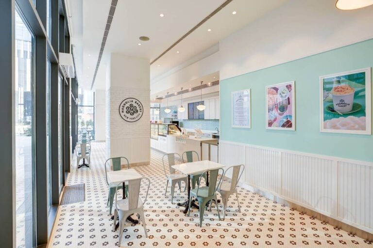 Bright cafe interior with mint-green wall, framed posters, white tables and metal chairs, and patterned tile floor by large windows.