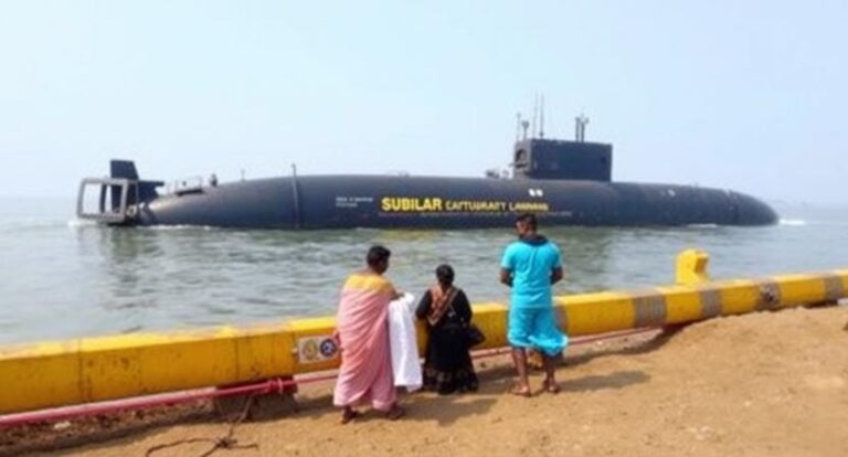 A large black submarine near a yellow seawall, with people standing on the shore watching it and some with towels or clothing draped over their shoulders.
