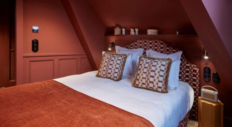 Cozy hotel bedroom with a large bed, white sheets, and red-brown bedding and pillows in a sloped-ceiling room.