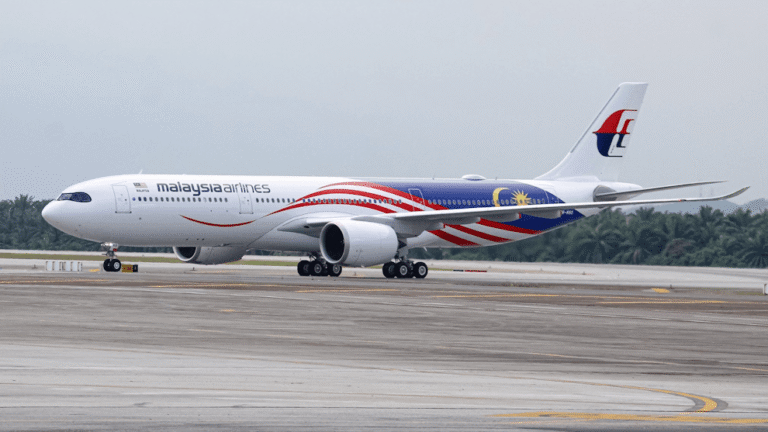Malaysia Airlines aircraft on a taxiway, showing red-blue flag livery and tail logo under a cloudy sky.