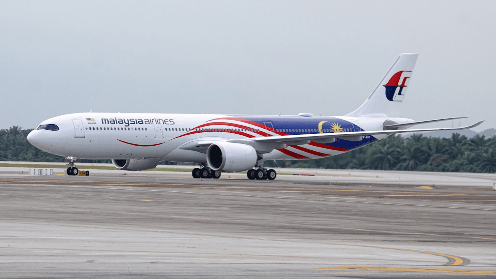Malaysia Airlines aircraft on a taxiway, showing red-blue flag livery and tail logo under a cloudy sky.