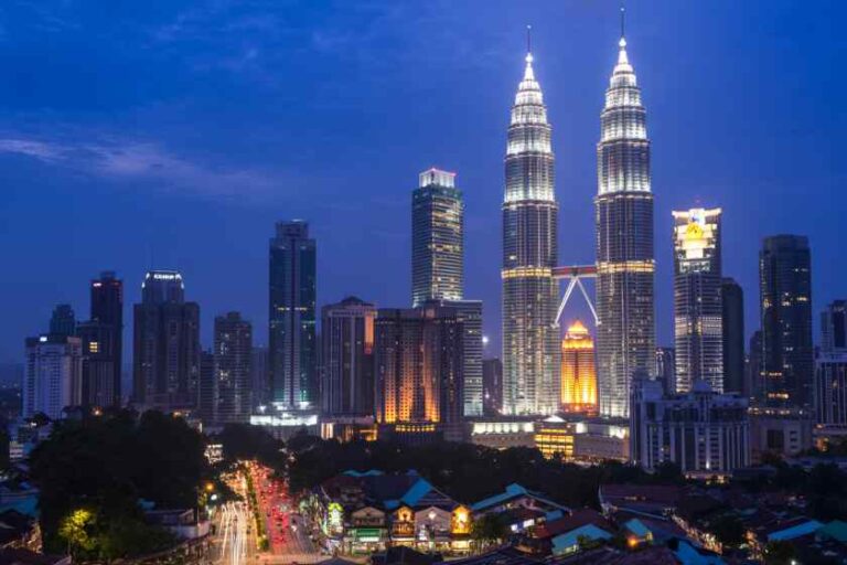 Nighttime cityscape of Kuala Lumpur with the Petronas Twin Towers glowing at the center among modern skyscrapers and a busy street below.