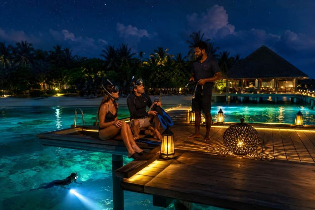 Nighttime scene at a tropical resort: three people on a wooden dock in snorkeling gear, with a swimmer below the platform and lanterns lighting the water edge that glows turquoise at dusk/night.