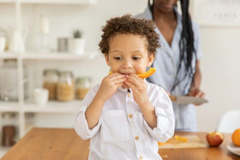 Young child eats an orange slice at a bright kitchen table, with a caregiver in the background.