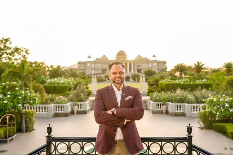 Man in a maroon blazer crosses his arms in a formal garden with a grand building in the background.