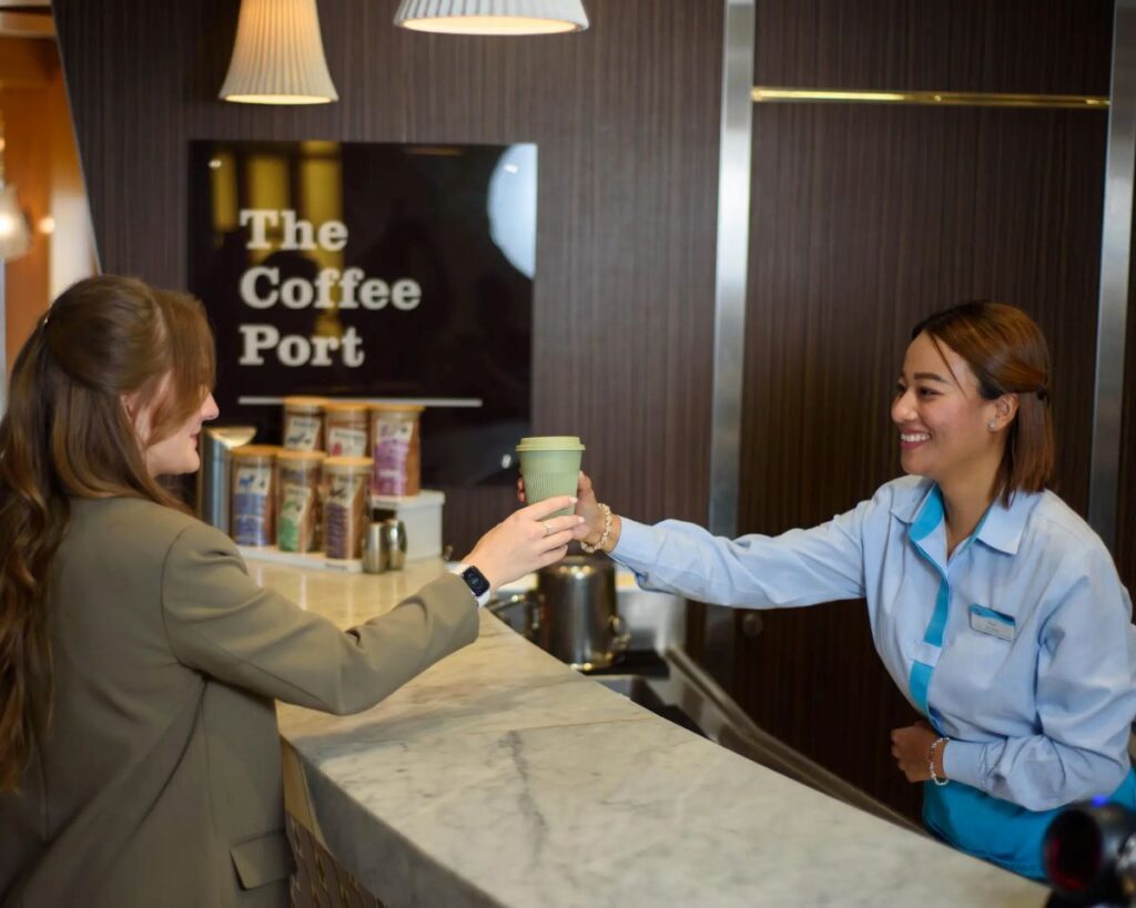 Customer hands a green disposable coffee cup to a smiling barista at a marble counter, The Coffee Port sign in background