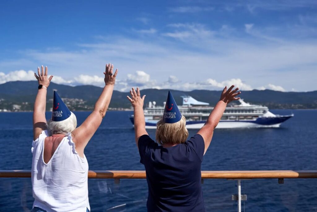 Two passengers on a cruise ship deck wearing Marella Cruises party hats raise their arms in celebration as a large cruise liner passes by on blue water with distant mountains.