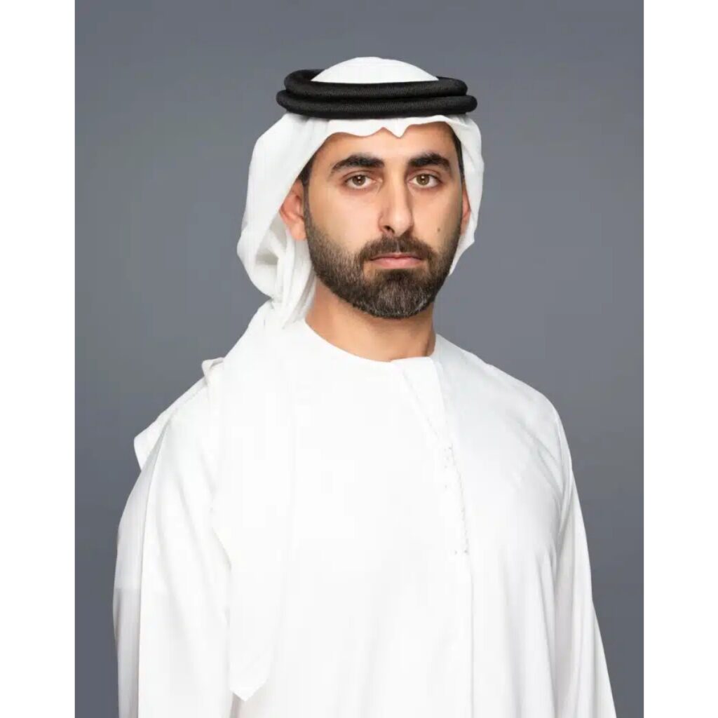 Portrait of a man with a beard wearing a white kandura and ghutra with a black agal against a gray background, looking at the camera.
