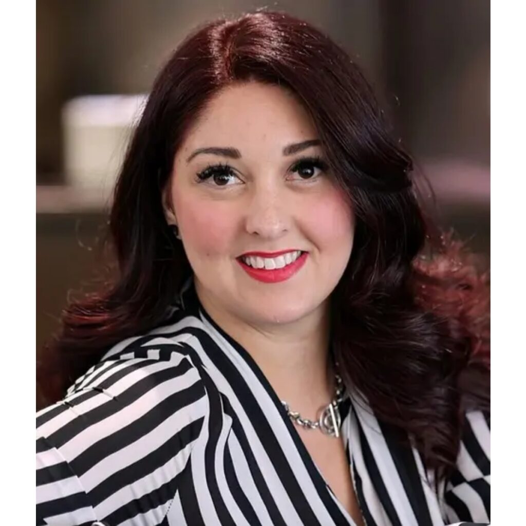 Portrait of a smiling woman with dark red hair, wearing a black-and-white striped blazer and a silver necklace.