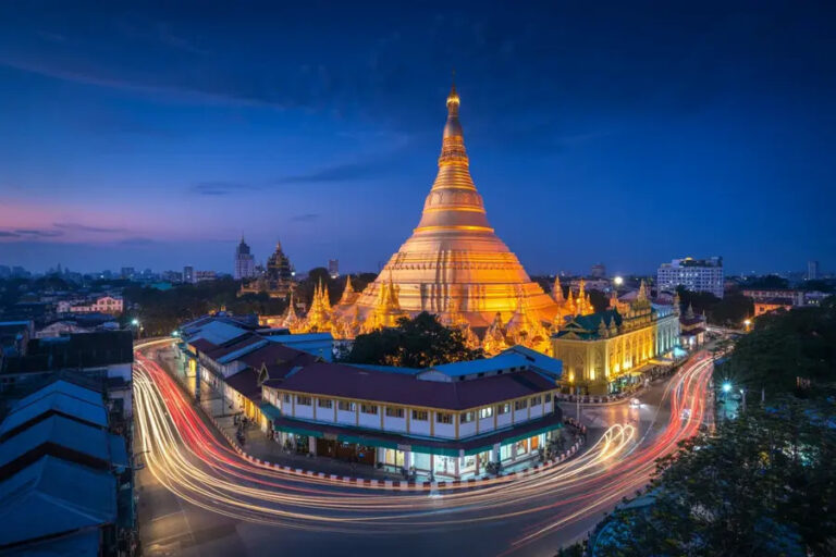 Golden illuminated stupa with surrounding smaller temples at dusk, city buildings in the background and light-trail streets.