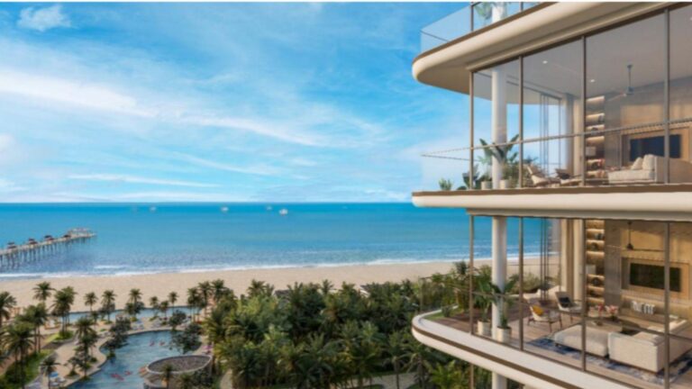 Modern glass-walled condo balcony overlooking a tropical beach and calm blue ocean, with palm trees below.